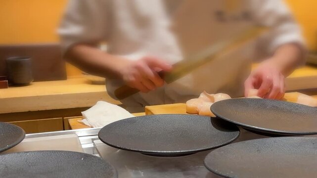 Professional sushi chef prepares fresh raw scallops at an authentic omakase restaurant counter in Tokyo. The master carefully cuts sea mollusk meat using a traditional long Japanese yanagiba knife.