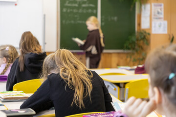 Elementary school students sit in a classroom as one focused pupil stands to read aloud from a textbook in front of the chalkboard during a collective academic learning session © Alina