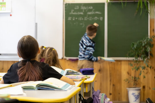 A primary school child solves math problems on a green blackboard in a classroom as classmates sit at desks, highlighting academic growth and active learning in an educational setting