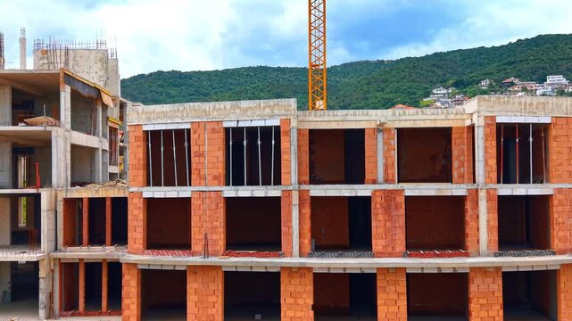 Concrete frame and brick walls of unfinished apartment building. Front view of a multi story residential complex during construction phase against green hills