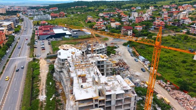 Aerial view of reinforced concrete building construction next to highway. Drone shot of monolithic concrete structure under development with yellow tower crane near road and green hills.