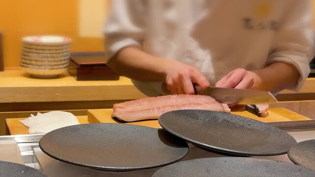 Professional Japanese sushi master slicing fresh raw tuna fish fillet on wooden cutting board in Tokyo. Experienced chef using authentic single bevel yanagiba knife to make cuts for premium sashimi.