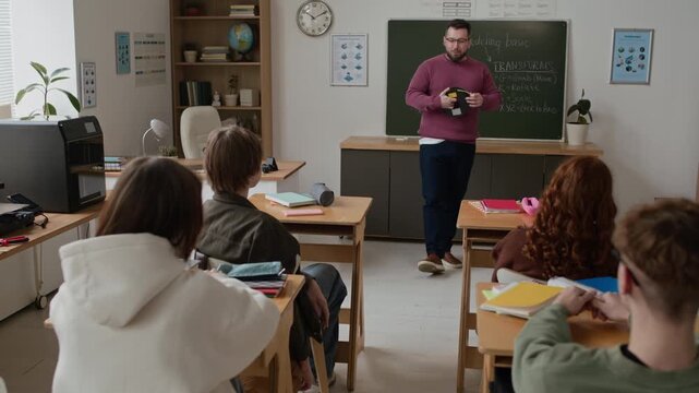 Male teacher showing students spool of filament during class in modern classroom equipped with 3D printer and a chalkboard