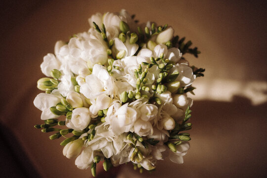 Overhead close-up of a round bouquet of white freesias with green buds, held in warm natural light that casts soft shadows on a beige background, elegant romantic bridal vibe