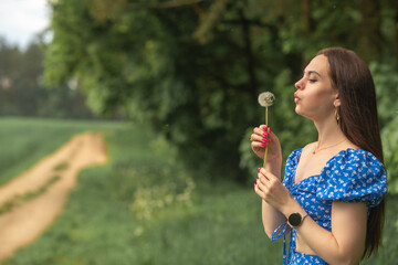 Fototapeta premium Young woman with long brown hair in blue floral dress blowing dandelion seeds in a green field, soft focus background with pathway and trees visible
