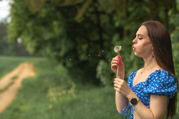 Fototapeta premium Young woman with long brown hair in blue floral dress blowing dandelion seeds in a green field, soft focus background with pathway and trees visible