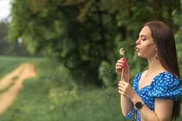 Fototapeta premium Young woman with long brown hair in blue floral dress blowing dandelion seeds in a green field, soft focus background with pathway and trees visible