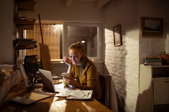 Smiling young woman using smartphone while working on laptop at cozy home office at night
