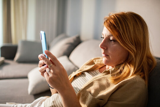 Red-haired woman using smartphone on sofa at home