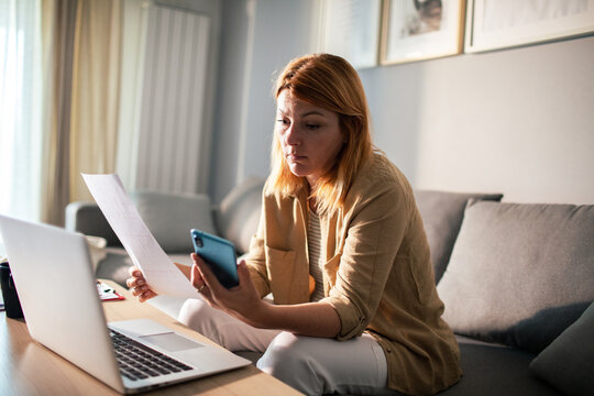 Concerned woman managing bills on smartphone with laptop at home living room