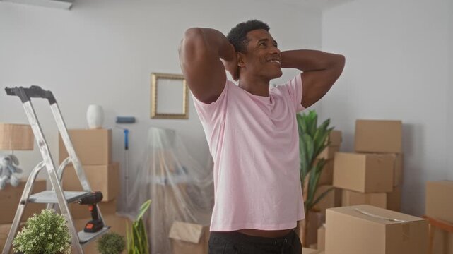Man with hands behind head, smiling while stretching among packed moving boxes, ladder and potted plant inside a building; relief transition.