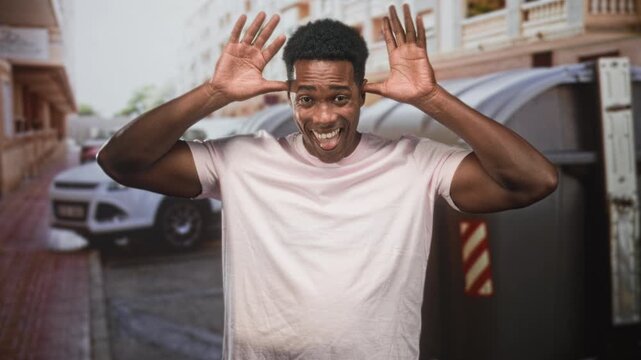 Man raising hands by head and smiling, wearing pink tshirt beside dumpster on street; playful energy mischief.