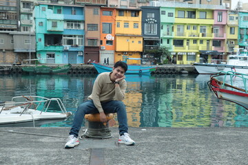 A man wearing a brown sweater and jeans stands posing by the pier with a view of colorful buildings in the background. © Peeb Kanom