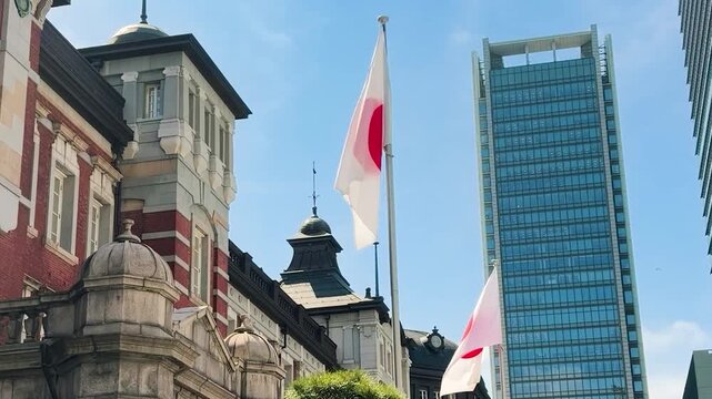 Japan flag waving near the historic Tokyo Station building in Marunouchi district. Red brick facade of the landmark railway station stands next to modern glass skyscrapers.