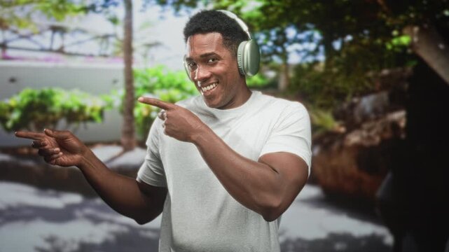 Man with headphones wearing white tee smiling broadly points finger to right at building entrance with trees and shrubs; joy upbeat energy.