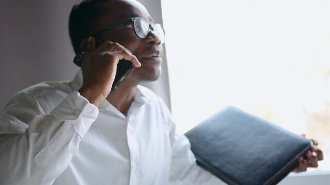 Black man in office talks on smartphone with humor and smiles while explaining views using a black leather folder for documents in hands, illustrating negotiation and communication skills