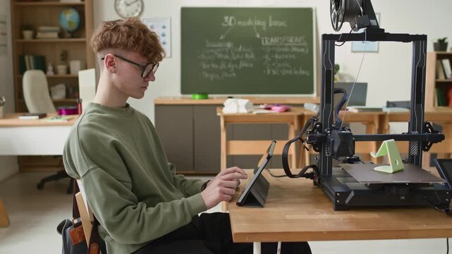 Student using digital tablet to control 3D printer, printing green smartphone stand, in modern classroom