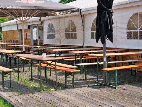 Rows of empty wooden beer garden tables and orange
benches stand on a rustic wooden terrace outside a large
festival tent. Large sun umbrellas, including one branded
with "Espiritu Libre"