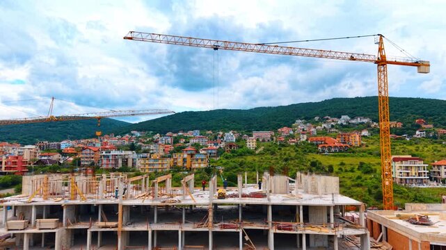 Multi story building construction site with yellow tower cranes. Wide shot of concrete residential building under construction with cranes against a hillside town and cloudy sky.