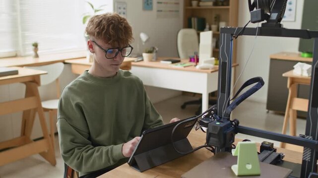 Student, wearing glasses and green sweatshirt, sitting at table, and looking at digital tablet connected to 3D printer in classroom