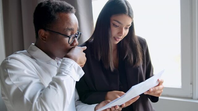 Black man and Indian woman colleagues review paper documents together at a modern office during the day. They discuss ideas, point to content, and react with smiles and excitement