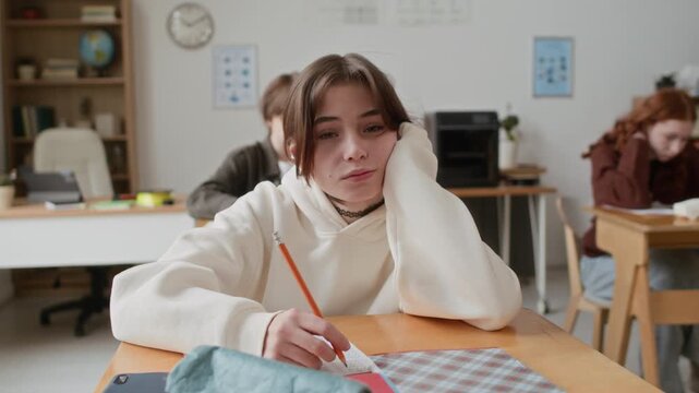 Portrait shot of bored teenage girl in hoodie lying on her desk and resting her head on her hand while looking at camera in modern classroom