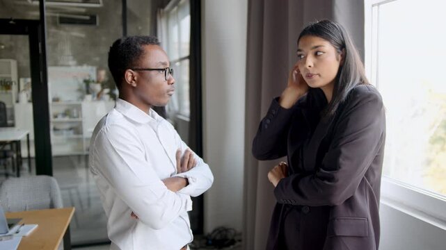 Two diverse colleagues a Black man and an Indian woman communicate in an office. They are engaged in a serious discussion during a workday break standing by a bright window