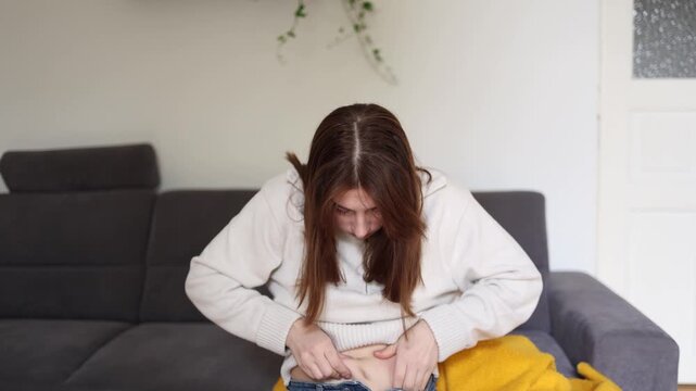 Young woman disappointed with slow weight loss progress touches stomach at home feeling insecure about body shape and summer appearance after fitness efforts