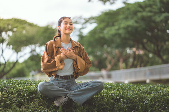 Peaceful young Asian woman sitting on grass with closed eyes and hands on chest, practicing mindfulness and deep breathing in summer park, happy female relaxing outdoors, mental health and wellness