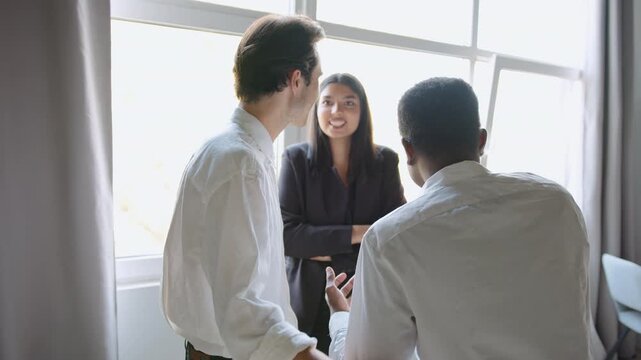 Three diverse professional colleagues, two men and one woman, conversing cheerfully during a daytime break in a bright office. They stand near a window, sharing a relaxed moment and smiling