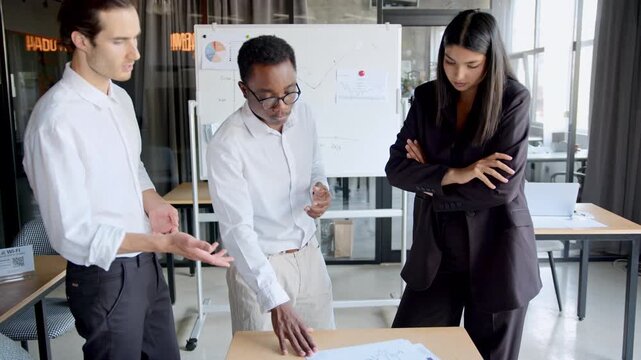 An experienced Black mentor explains financial charts and reports to two diverse young colleagues in a bright office. They discuss data analysis methods to improve business performance and trade