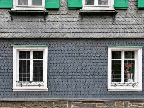 A symmetrical straight shot captures a traditional German
house facade covered in dark gray slate shingles. The
historic architecture features white latticed windows with
decorative wrought iron grill