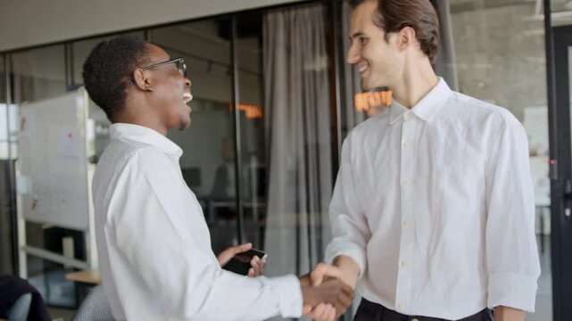 Smiling Black manager warmly greets a new White male employee and an Indian female employee with handshakes in a modern office, anticipating strong teamwork and a good working atmosphere.