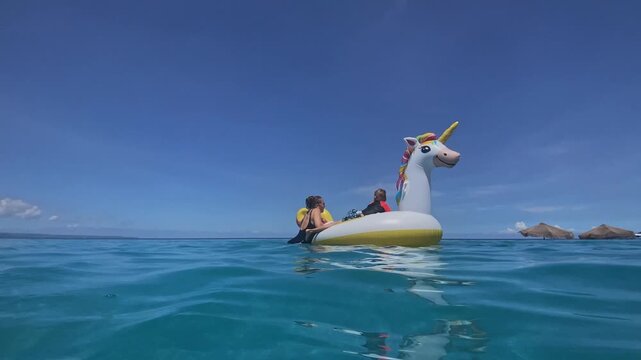 Mother and son on unicorn float, sunlit turquoise sea, gentle waves lapping, island horizon, giggling child, affectionate parenting, summer vacation mood, playful splashes, candid, Bali, Indonesia