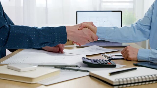 Business partners shaking hands over architectural project blueprints at office desk. Professional men making deal or successful construction contract agreement near laptop.