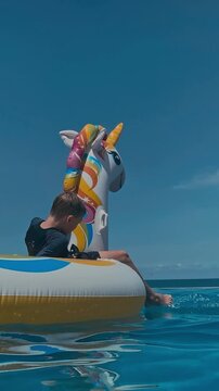 Inflatable unicorn floating on ocean water, boys clinging to ring in snorkel gear under clear blue sky, gentle waves reflecting sunlight, playful summer vacation mood, lowangle, vertical video