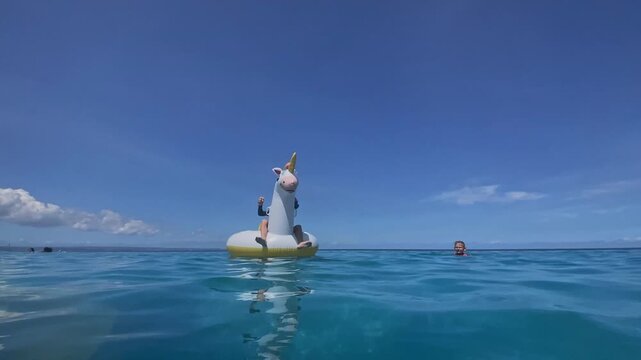 Caucasian boy on unicorn float drifting across calm turquoise ocean under clear blue sky, reflective water surface, distant clouds, slow ripples, relaxed summer leisure, Bali island