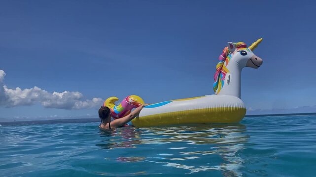 Woman on inflatable unicorn float ocean, blue sky and turquoise water, female tourist clinging to colorful float, gentle waves, sunlit summer vacation vibe with playful splashes and laughter,Bali