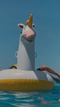 Closeup inflatable unicorn float with boy wearing goggles, turquoise pool water shimmering, sunny sky, playful young snorkeler gripping rim, summer joy and carefree motion, vertical video