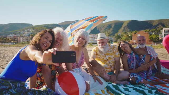 Group of cheerful senior friends lying on beach towels, smiling and laughing together at the camera during a beautiful sunny day, representing a concept of happy retirement and friendship