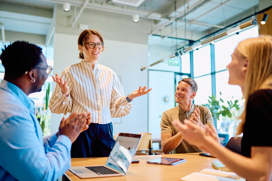 Business executive presenting to colleagues in a modern office meeting, team members are clapping, showing appreciation and success