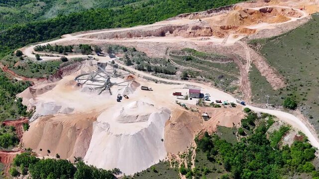 Aerial view of an open-pit quarry and stone mining terraces surrounded by green hills. Industrial mining landscape against green forests and blue sky.	
