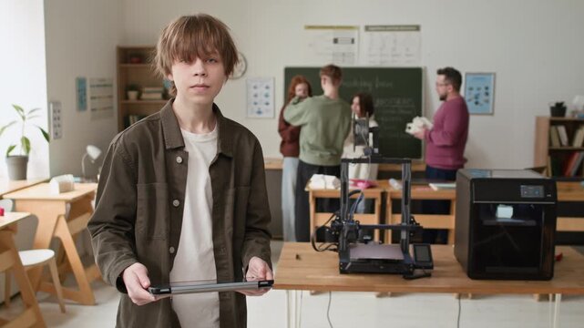 Portrait shot of student holding tablet, looking up at camera against two 3D printers and group of students engaged in discussion in classroom