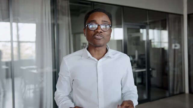 Black male professional wearing glasses and a white shirt presents his groundbreaking startup project with animated gestures at a busy office meeting during business hours