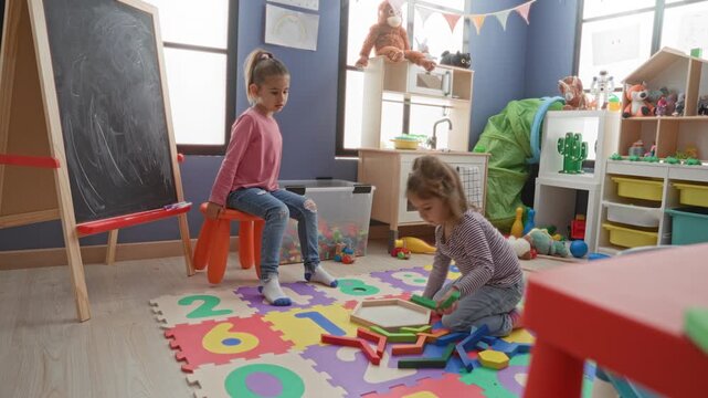 Girl seated on an orange stool beside a chalkboard while another child kneels on a foam number mat arranging wooden blocks in a playroom building; curiosity learning playful.