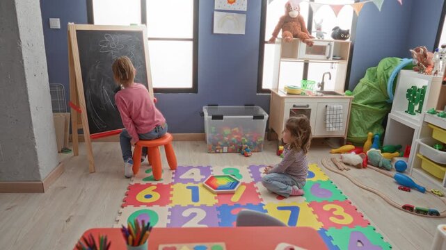 Girl draws on chalkboard while another child kneels on a number puzzle mat and stacks toys in an indoor playroom building; playful learning.