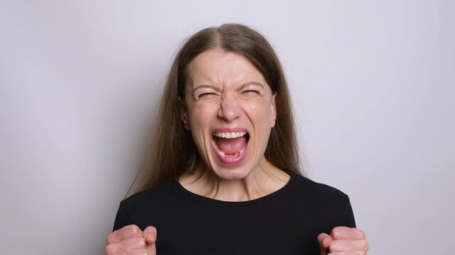 Furious woman screaming with wide open mouth and clenched fists against grey background. Angry female expressing rage, hatred or intense aggression during nervous breakdown.