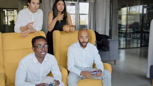 Diverse colleagues enjoy a lively console game break in a modern office, fostering unity and celebrating an exciting team victory with joysticks