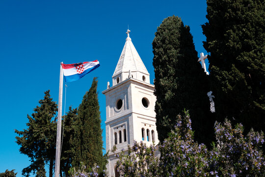 A white stone bell tower stands tall beside a waving Croatian flag and green cypress trees on a sunny day in Primosten, Croatia