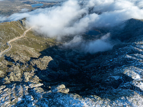 Aerial view of the rugged peaks of Serra da Estrela with thick clouds drifting over a winding mountain road and a blue glacial lake Unhais da Serra, Castelo Branco, Portugal.